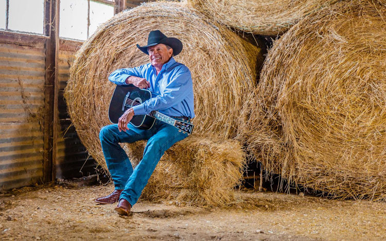 George Strait sitting on a square bale holding a guitar smiling.